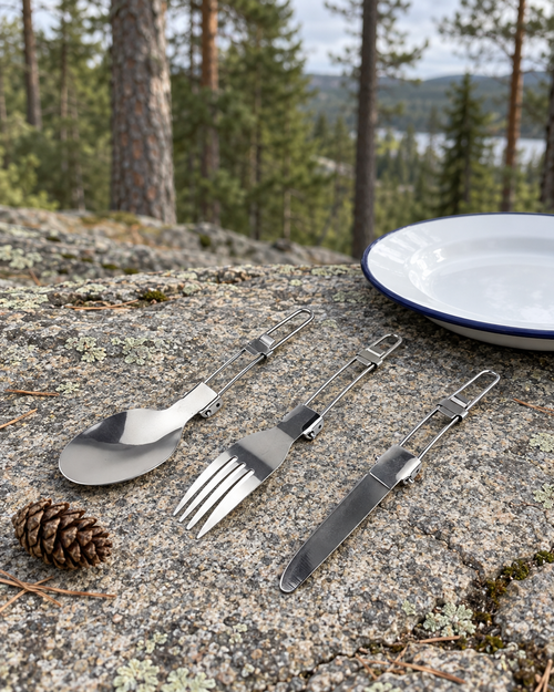 Folding stainless steel spoon, fork and table knife on a granite ledge beside a white enamel camp plate, a pine cone for scale