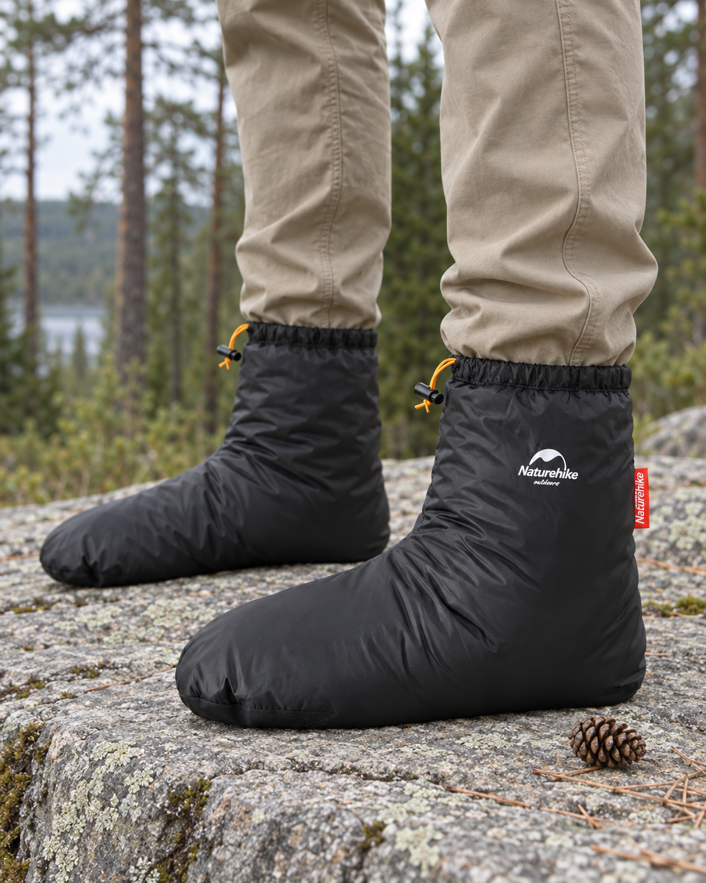 Pair of black down booties worn over beige trousers on a lichen-covered granite ledge at the edge of a pine forest, drawcords cinched at the ankles