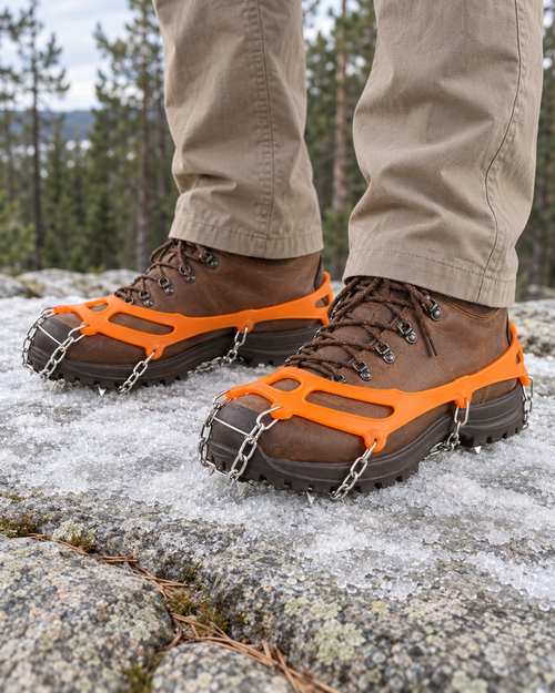 Orange chain crampons worn over brown leather hiking boots on icy granite, steel chains and spikes under the soles