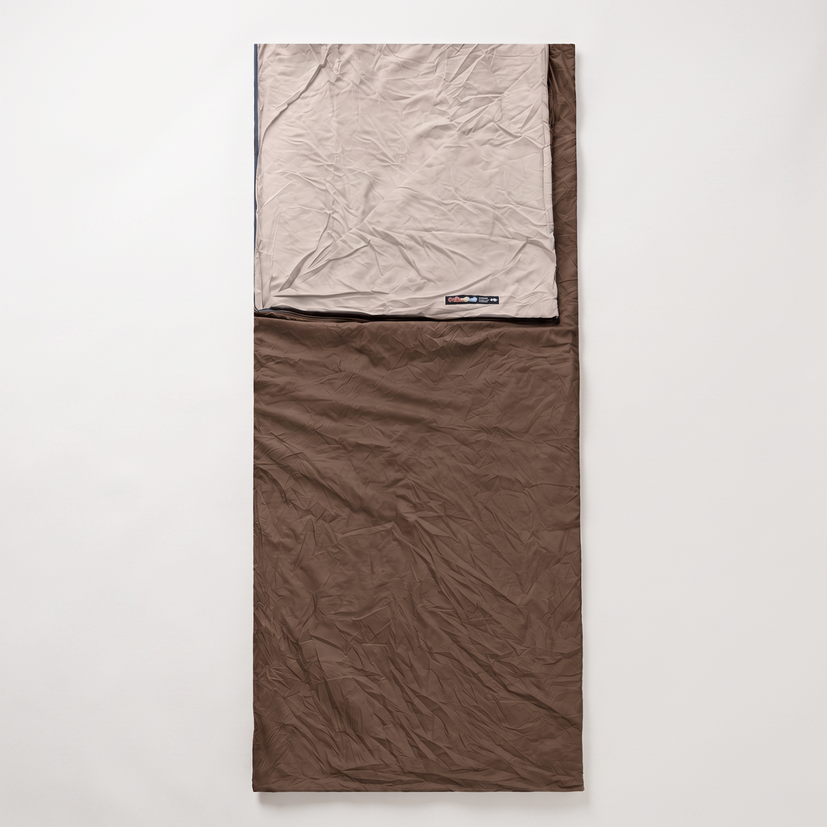 A brown envelope sleeping bag laid flat on a pale background, its cream lining folded back at the head above a small woven label