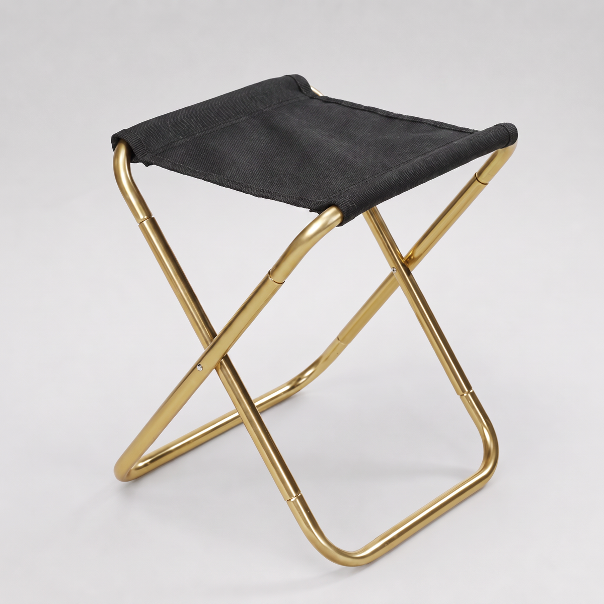Compact folding camp stool with a gold aluminium X-frame and a black woven fabric seat on a seamless pale grey paper sweep.