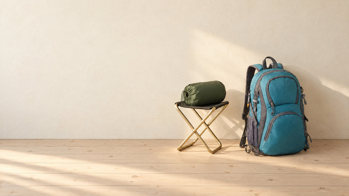 A folding camping stool with a packed sleeping bag and a teal hiking backpack on pale pine floorboards in morning sun